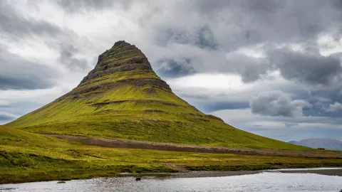 Mountain with green slopes under cloudy sky, reflected in water.