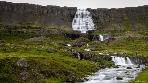 Waterfall cascading down rocky cliffs, surrounded by lush green landscape.