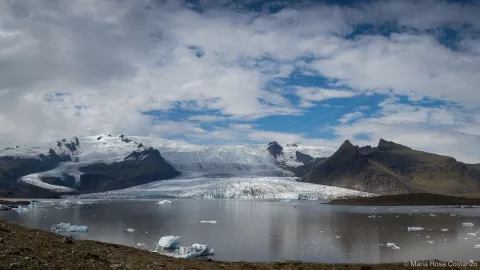 Glacier with floating ice on a calm lake, under a cloudy sky.