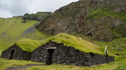 Grass-covered stone huts at the base of a green, rocky hillside.